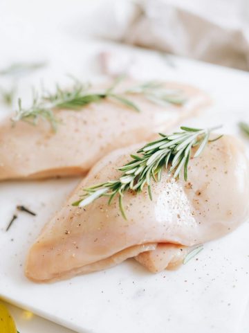 Close-up of raw chicken breasts garnished with fresh rosemary and spices on a cutting board.